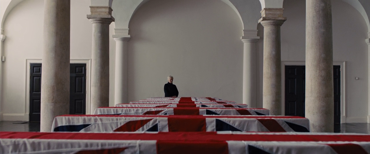 A screenshot from the movie 'Skyfall'. The character 'M' looks over the a line of coffins draped with Union flags.